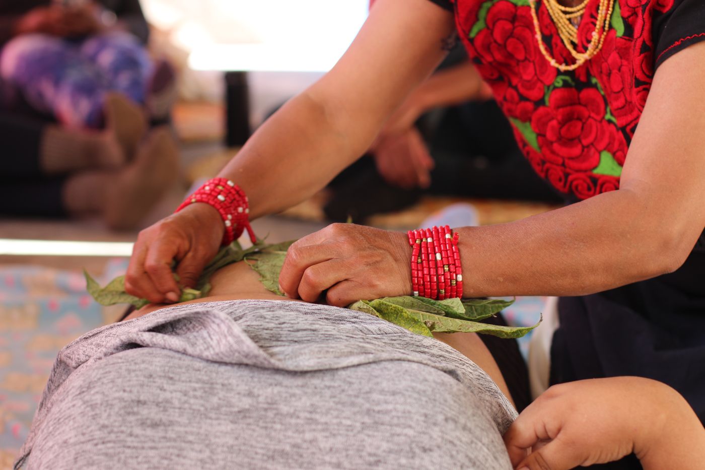 Hands with red beaded bracelets placing fresh leaves on a woman's body during a treatment